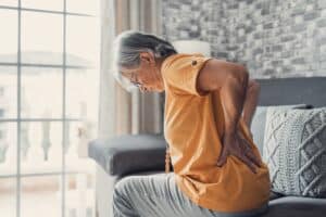Older woman sitting on bed holding back in pain from spinal stenosis in Houston, TX